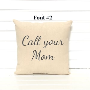 May include: A square, beige pillow with the text "Call your Mom" in a dark gray script font. The pillow has a simple design and is placed against a white background.
