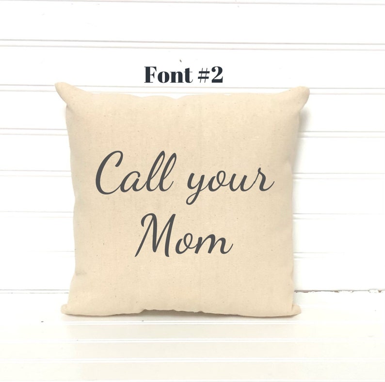 May include: A square, beige pillow with the text "Call your Mom" in a dark gray script font. The pillow has a simple design and is placed against a white background.