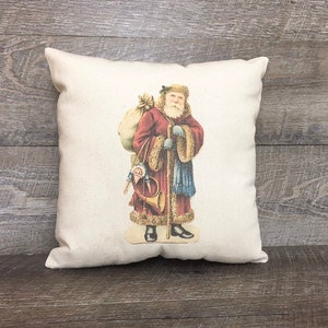 May include: A white pillow with a vintage illustration of Santa Claus carrying a bag of gifts and a horn. He is wearing a red and white suit with a blue sash and a white fur trim.