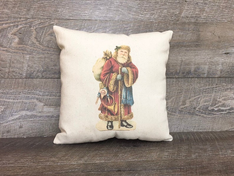 May include: A white pillow with a vintage illustration of Santa Claus carrying a bag of gifts and a horn. He is wearing a red and white suit with a blue sash and a white fur trim.