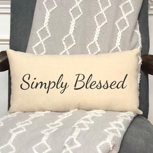 May include: A beige throw pillow with the text "Simply Blessed" in black cursive font. The pillow is on a gray chair with a white and gray patterned throw blanket.