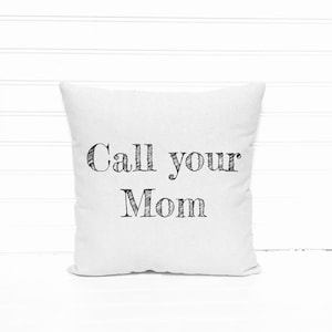 May include: Square white pillow with the text "Call your Mom" in black, handwritten-style lettering. The pillow is centered against a white, horizontal-slat background.