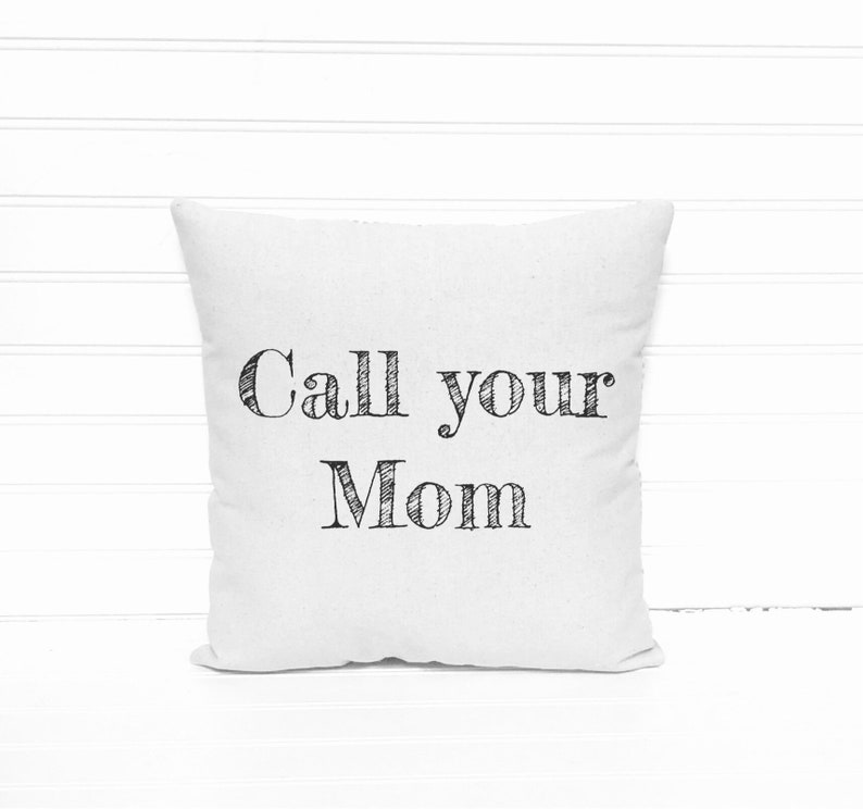May include: Square white pillow with the text "Call your Mom" in black, handwritten-style lettering. The pillow is centered against a white, horizontal-slat background.