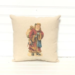 May include: A cream-colored pillow with a vintage illustration of Santa Claus carrying a sack of gifts and a horn. He is wearing a red coat with white fur trim and a blue scarf.
