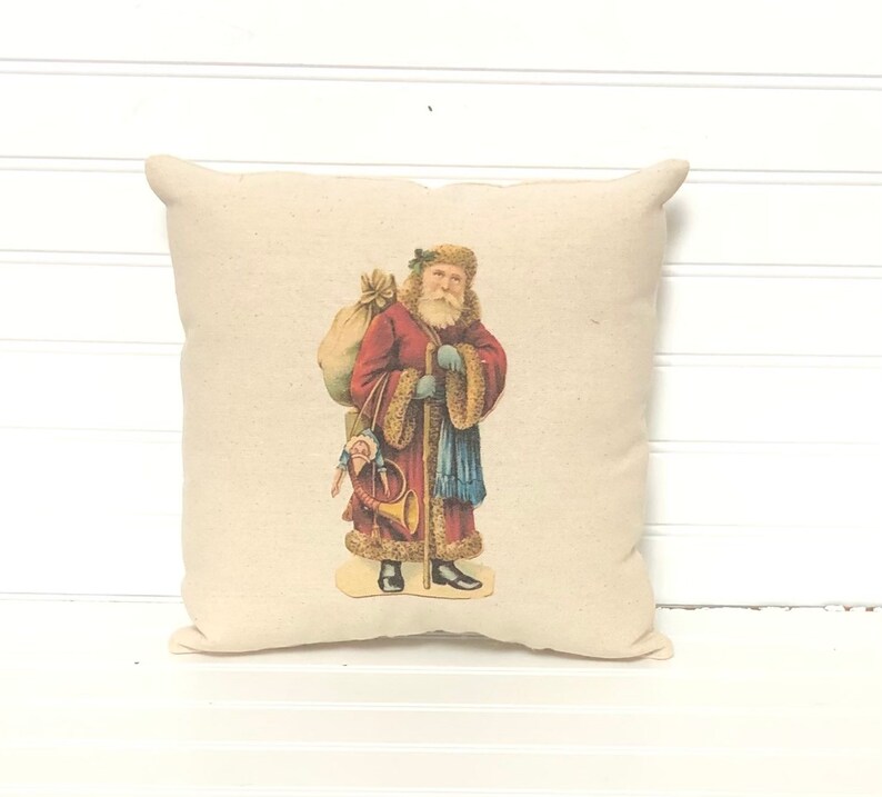 May include: A cream-colored pillow with a vintage illustration of Santa Claus carrying a sack of gifts and a horn. He is wearing a red coat with white fur trim and a blue scarf.