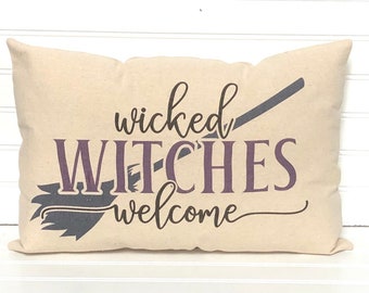 May include: Beige rectangular pillow with the words "wicked WITCHES welcome" in purple and black lettering. A black witch's broom is also featured on the pillow. The pillow is a Halloween-themed decorative item.