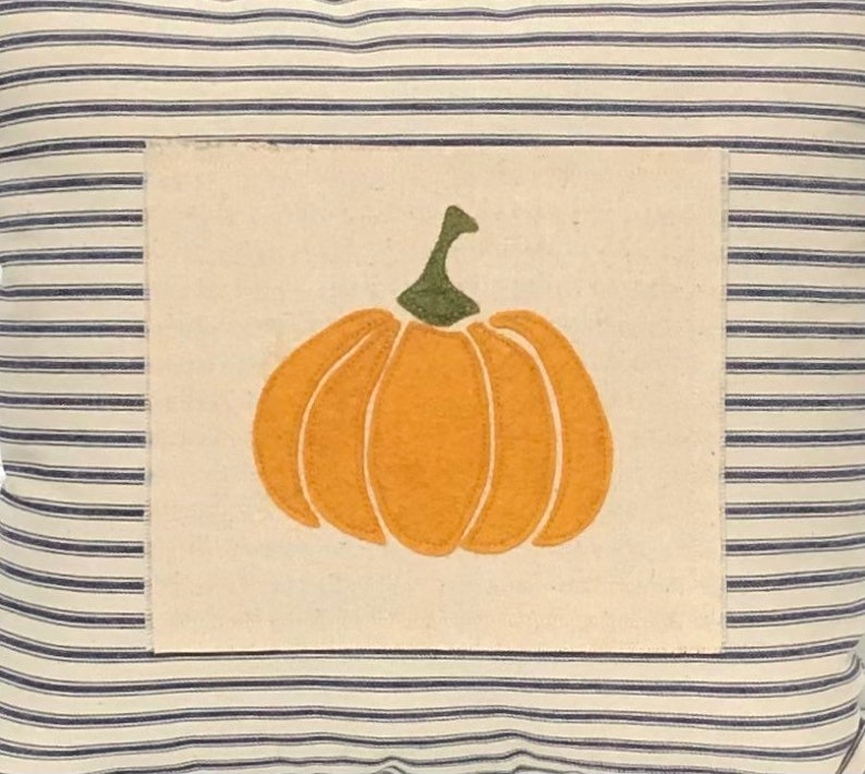 Puede incluir: Un coj&iacute;n cuadrado con una base de tela color crema y finas rayas horizontales azul marino. Centrado en el coj&iacute;n hay un parche rectangular de tela color blanco roto que muestra una aplicaci&oacute;n de calabaza naranja con un tallo verde oscuro.