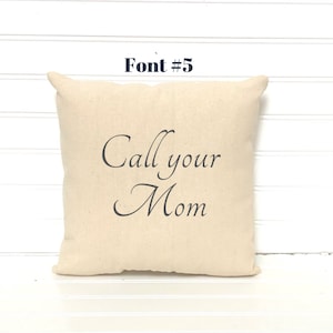 May include: A square, beige throw pillow with the phrase "Call your Mom" printed in elegant, dark gray script. The text "Font #5" is above the pillow. The pillow has a simple, classic design.