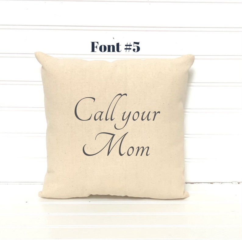 May include: A square, beige throw pillow with the phrase "Call your Mom" printed in elegant, dark gray script. The text "Font #5" is above the pillow. The pillow has a simple, classic design.