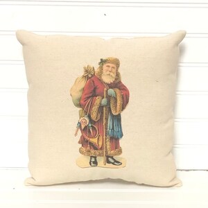 May include: A cream-colored pillow with a vintage illustration of Santa Claus carrying a bag of gifts and a horn. He is wearing a red coat with white fur trim and a blue scarf.