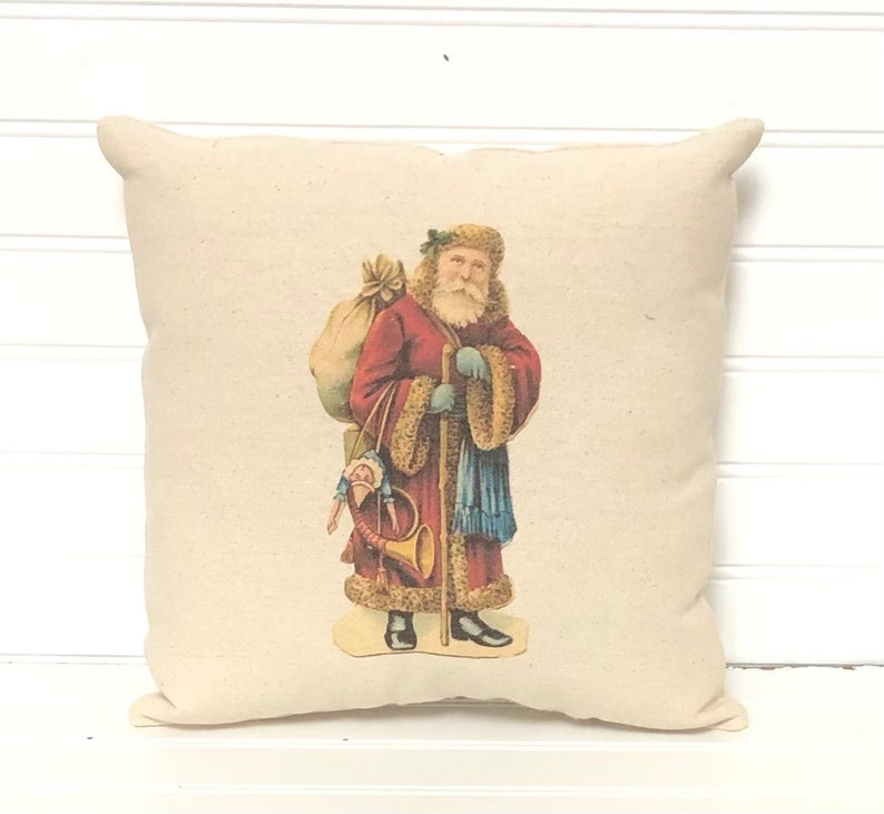 May include: A cream-colored pillow with a vintage illustration of Santa Claus carrying a bag of gifts and a horn. He is wearing a red coat with white fur trim and a blue scarf.