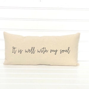 May include: A rectangular beige pillow with the phrase "It is well with my soul" printed in black script. The pillow is placed on a white surface with horizontal lines.