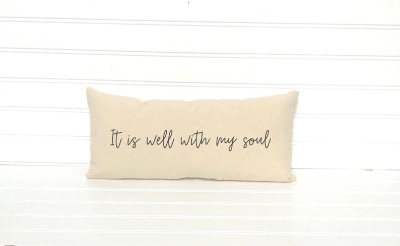 May include: A rectangular beige pillow with the phrase "It is well with my soul" printed in black script. The pillow is placed on a white surface with horizontal lines.