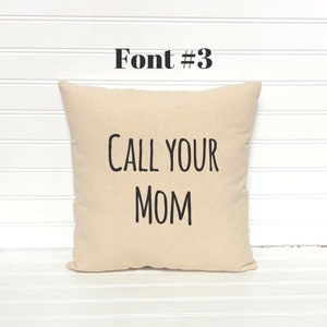 May include: A beige square pillow with the text "CALL YOUR MOM" in black, bold font. Above the pillow, the text "Font #3" is also in black. The pillow is on a white, slatted surface.