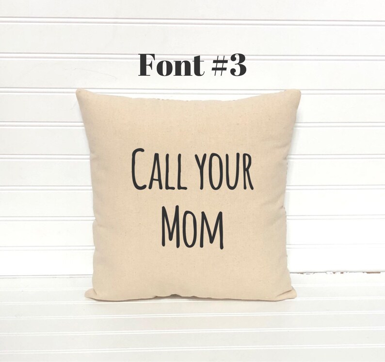 May include: A beige square pillow with the text "CALL YOUR MOM" in black, bold font. Above the pillow, the text "Font #3" is also in black. The pillow is on a white, slatted surface.