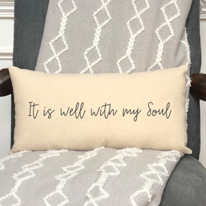 May include: Cream-colored rectangular pillow with the text "It is well with my Soul" in black script. The pillow is placed on a gray chair with a patterned throw blanket.