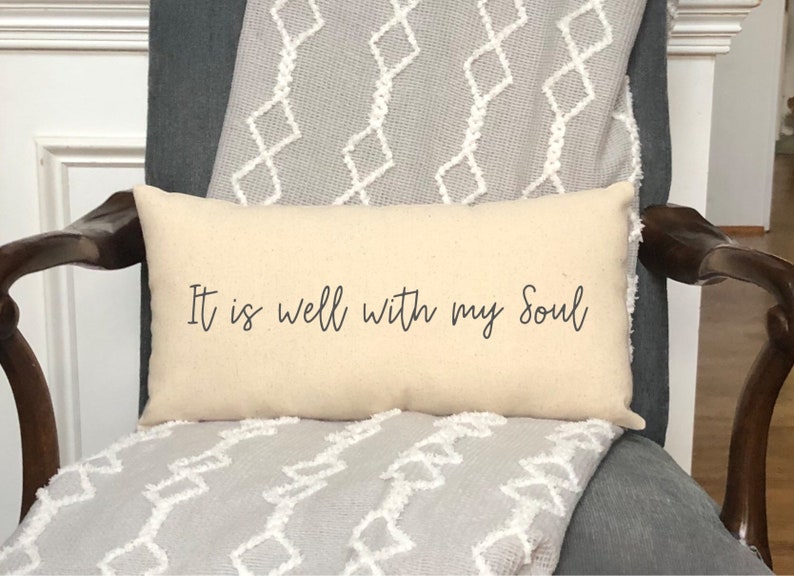 May include: Cream-colored rectangular pillow with the text "It is well with my Soul" in black script. The pillow is placed on a gray chair with a patterned throw blanket.