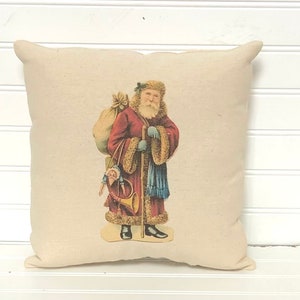 May include: A cream-colored pillow with a vintage illustration of Santa Claus carrying a bag of toys and a horn. Santa is wearing a red coat with fur trim, blue pants, and a white beard.