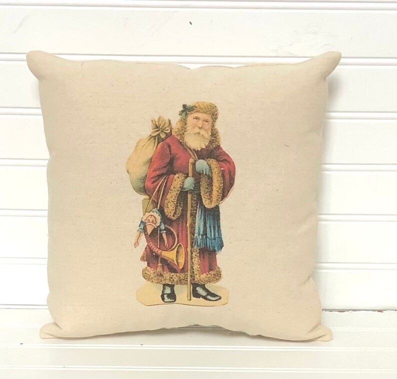 May include: A cream-colored pillow with a vintage illustration of Santa Claus carrying a bag of toys and a horn. Santa is wearing a red coat with fur trim, blue pants, and a white beard.