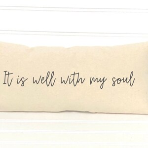 May include: A rectangular, cream-colored pillow with the phrase "It is well with my soul" printed in black script. The pillow is placed against a white, horizontal-slat background.