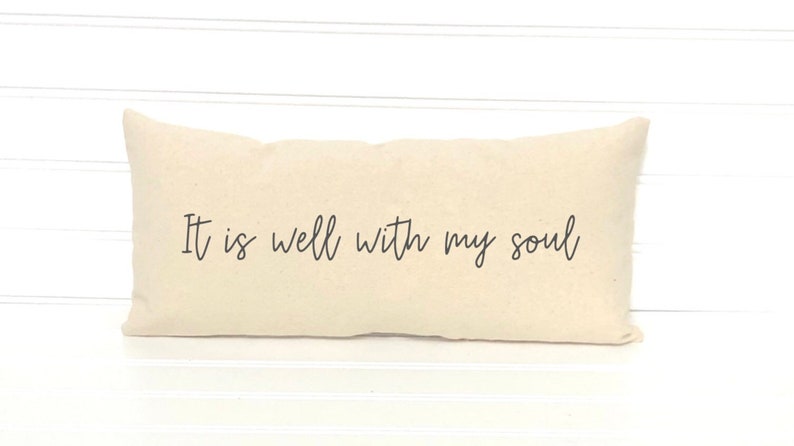 May include: A rectangular, cream-colored pillow with the phrase "It is well with my soul" printed in black script. The pillow is placed against a white, horizontal-slat background.