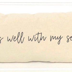 May include: Cream-colored rectangular pillow with the phrase "It is well with my soul" in black script. The pillow has a soft, textured appearance and is likely made of cotton or linen. A decorative item for a bed or sofa.