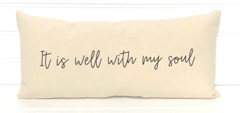 May include: Cream-colored rectangular pillow with the phrase "It is well with my soul" in black script. The pillow has a soft, textured appearance and is likely made of cotton or linen. A decorative item for a bed or sofa.