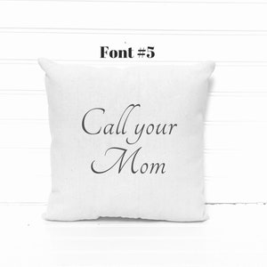 May include: A square, off-white pillow with the phrase "Call your Mom" printed in elegant black script. The text is centered on the pillow, which has a soft, textured appearance. Above the pillow is the text "Font #5" in a bold, black font.
