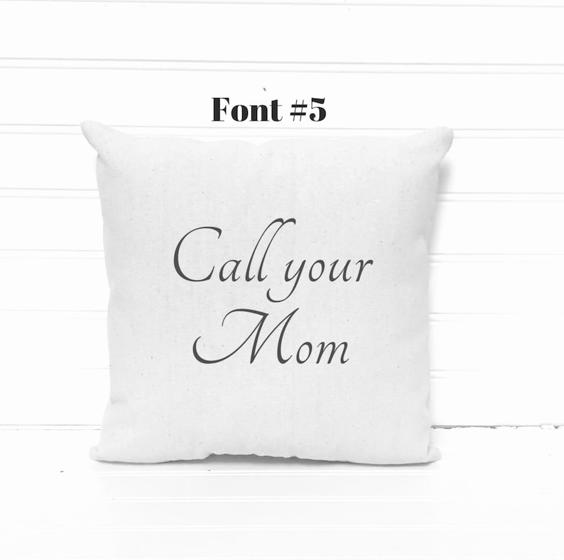 May include: A square, off-white pillow with the phrase "Call your Mom" printed in elegant black script. The text is centered on the pillow, which has a soft, textured appearance. Above the pillow is the text "Font #5" in a bold, black font.