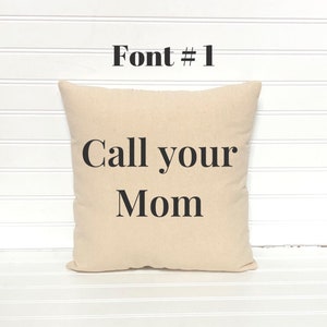 May include: A square beige pillow with the text "Call your Mom" in black, centered on the front. Above the text is the phrase "Font # 1" in black. The pillow is on a white slatted surface.