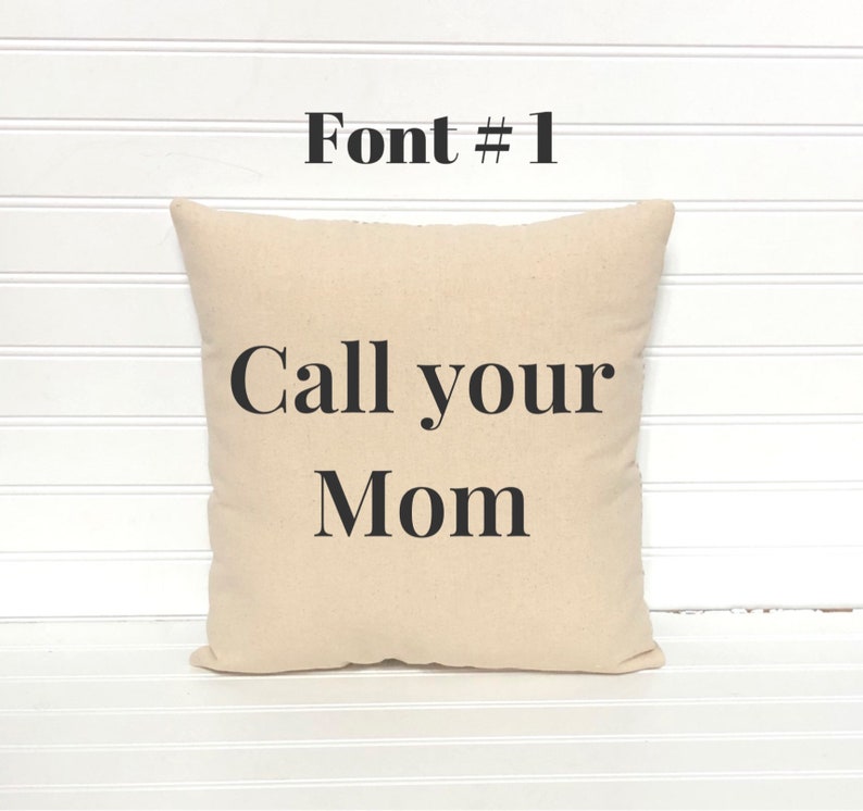 May include: A square beige pillow with the text "Call your Mom" in black, centered on the front. Above the text is the phrase "Font # 1" in black. The pillow is on a white slatted surface.