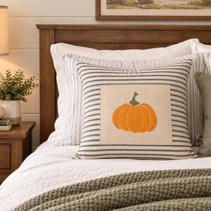 Puede incluir: Un coj&iacute;n cuadrado con un parche central de tela beige que presenta un dise&ntilde;o de calabaza naranja bordada. El coj&iacute;n tiene un patr&oacute;n de finas rayas horizontales de color azul oscuro sobre un fondo claro y est&aacute; acabado con un borde ribeteado.