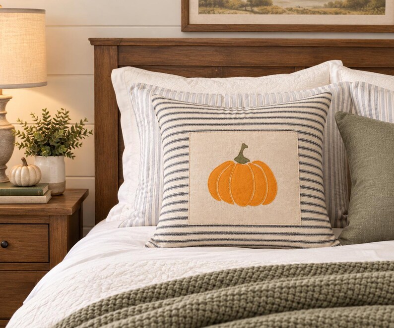 Puede incluir: Un coj&iacute;n cuadrado con un parche central de tela beige que presenta un dise&ntilde;o de calabaza naranja bordada. El coj&iacute;n tiene un patr&oacute;n de finas rayas horizontales de color azul oscuro sobre un fondo claro y est&aacute; acabado con un borde ribeteado.