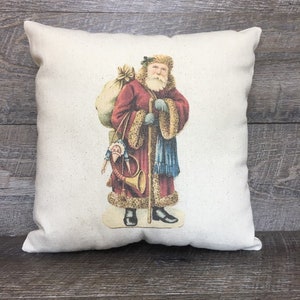 May include: A white pillow with a vintage illustration of Santa Claus carrying a bag of toys and a horn. He is wearing a red coat, a blue scarf, and a white fur trim.