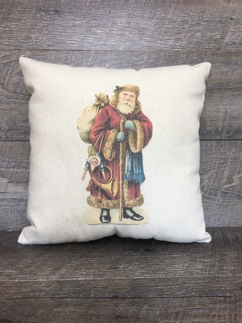 May include: A white pillow with a vintage illustration of Santa Claus carrying a bag of toys and a horn. He is wearing a red coat, a blue scarf, and a white fur trim.
