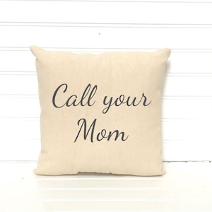May include: A square, beige pillow with the phrase "Call your Mom" printed in black script. The pillow has a soft, textured appearance and is likely made of cotton or a similar fabric. The pillow is a decorative item.