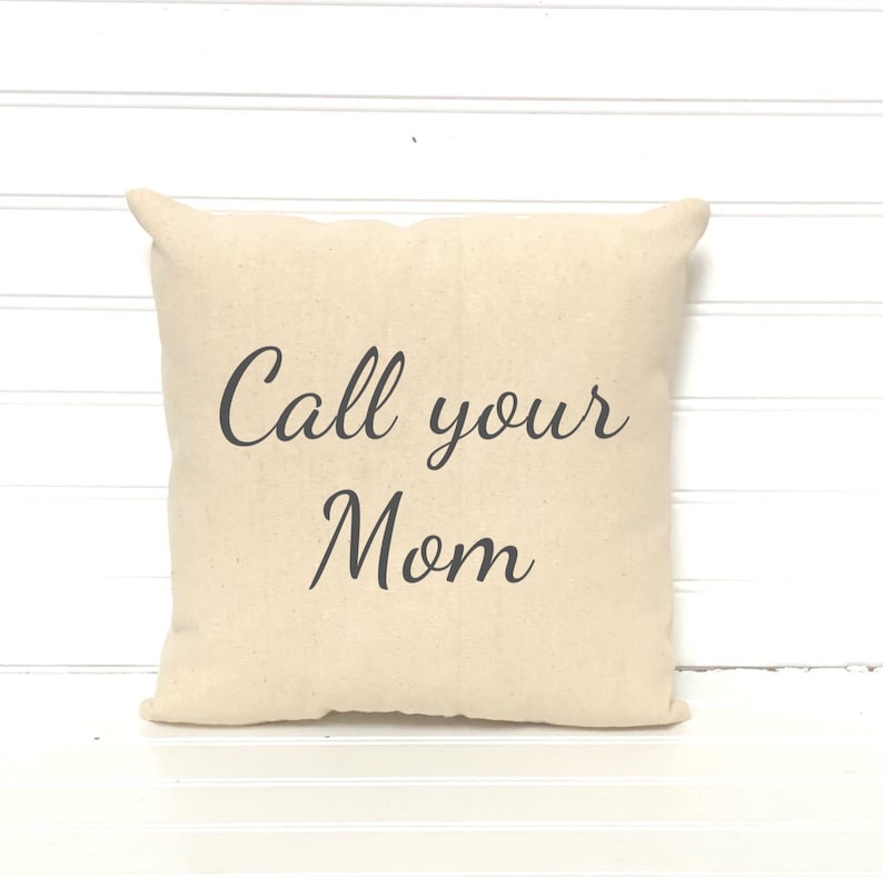 May include: A square, beige pillow with the phrase "Call your Mom" printed in black script. The pillow has a soft, textured appearance and is likely made of cotton or a similar fabric. The pillow is a decorative item.