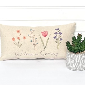 May include: A beige pillow with a floral design and the text "Welcome Spring". The pillow is decorated with a red tulip, pink and orange flowers, and purple flowers.