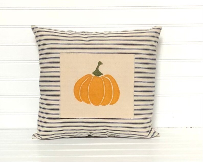 Puede incluir: Un coj&iacute;n cuadrado con una base de tela color crema y finas rayas horizontales azul marino. Centrado en la parte frontal hay un parche rectangular de tela color blanco roto que muestra una calabaza naranja con un tallo verde.