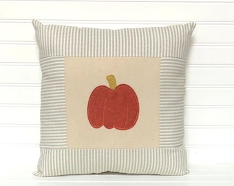 Puede incluir: Un cojín cuadrado que presenta un panel central beige con una calabaza de color naranja quemado aplicada y un tallo de color tostado. El borde circundante consiste en un tejido de rayas verticales finas en gris claro y blanco.