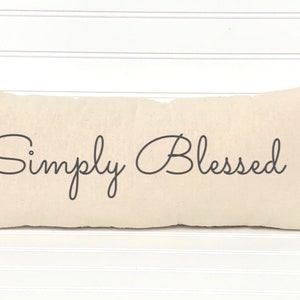 May include: A long, rectangular beige pillow with the text "Simply Blessed" printed in black cursive script.