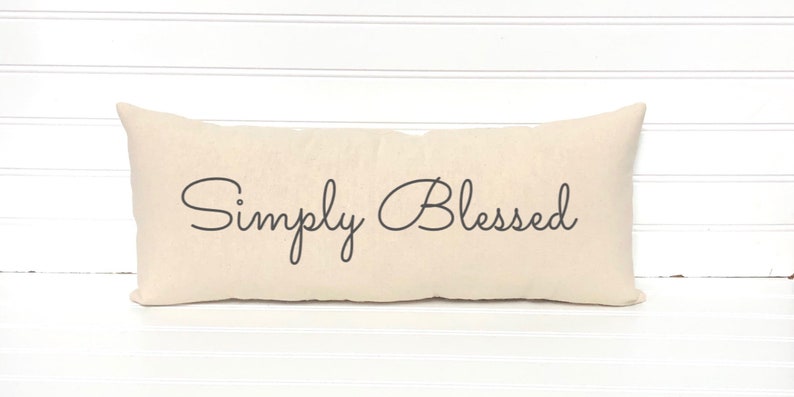May include: A long, rectangular beige pillow with the text "Simply Blessed" printed in black cursive script.