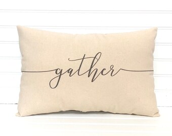 May include: A rectangular beige pillow with the word "gather" in a dark brown script font. The word is centered and underlined with a thin line. The pillow is against a white, slatted background.