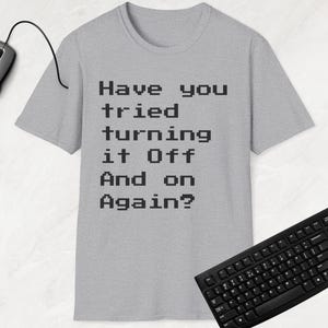 Op de afbeelding: Een grijs T-shirt met de tekst "Heb je geprobeerd het uit en weer aan te zetten?" in zwart gedrukt.