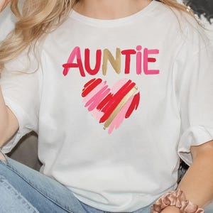 Peut inclure: T-shirt blanc avec le mot "AUNTIE" en lettres roses et rouges avec un motif de cœur doré.