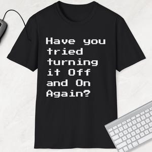 Op de afbeelding: Zwart T-shirt met witte tekst die "Heb je geprobeerd het uit en weer aan te zetten?" luidt. De tekst is in een gepixelde lettertype, doet denken aan oude videogames.