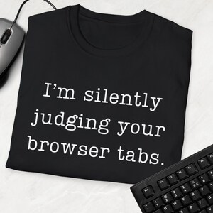 Op de afbeelding: Zwart T-shirt met witte tekst die "Ik beoordeel je browsertabbladen stilletjes." zegt.