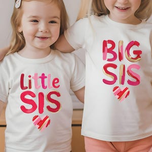 Peut inclure: Deux enfants portant des t-shirts blancs assortis. L'un dit "Little Sis" en rose et rouge avec un contour doré et un cœur. L'autre dit "Big Sis" dans le même style avec un cœur.