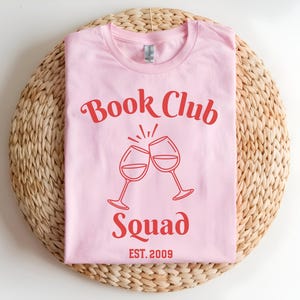 Op de afbeelding: Een lichtroze t-shirt met de rode tekst "Book Club Squad" en twee klinkende wijnglazen. De tekst "EST. 2009" staat eronder. Het shirt ligt op een ronde, geweven mat.