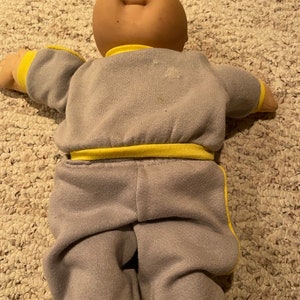 May include: A Cabbage Patch Kid doll wearing a gray tracksuit with yellow accents. The doll has a light brown complexion and a bald head.